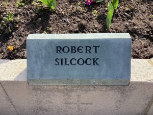 A grey slate stone mounted on top of a low wall surrounding a garden. It bears the name ROBERT SILCOCK.