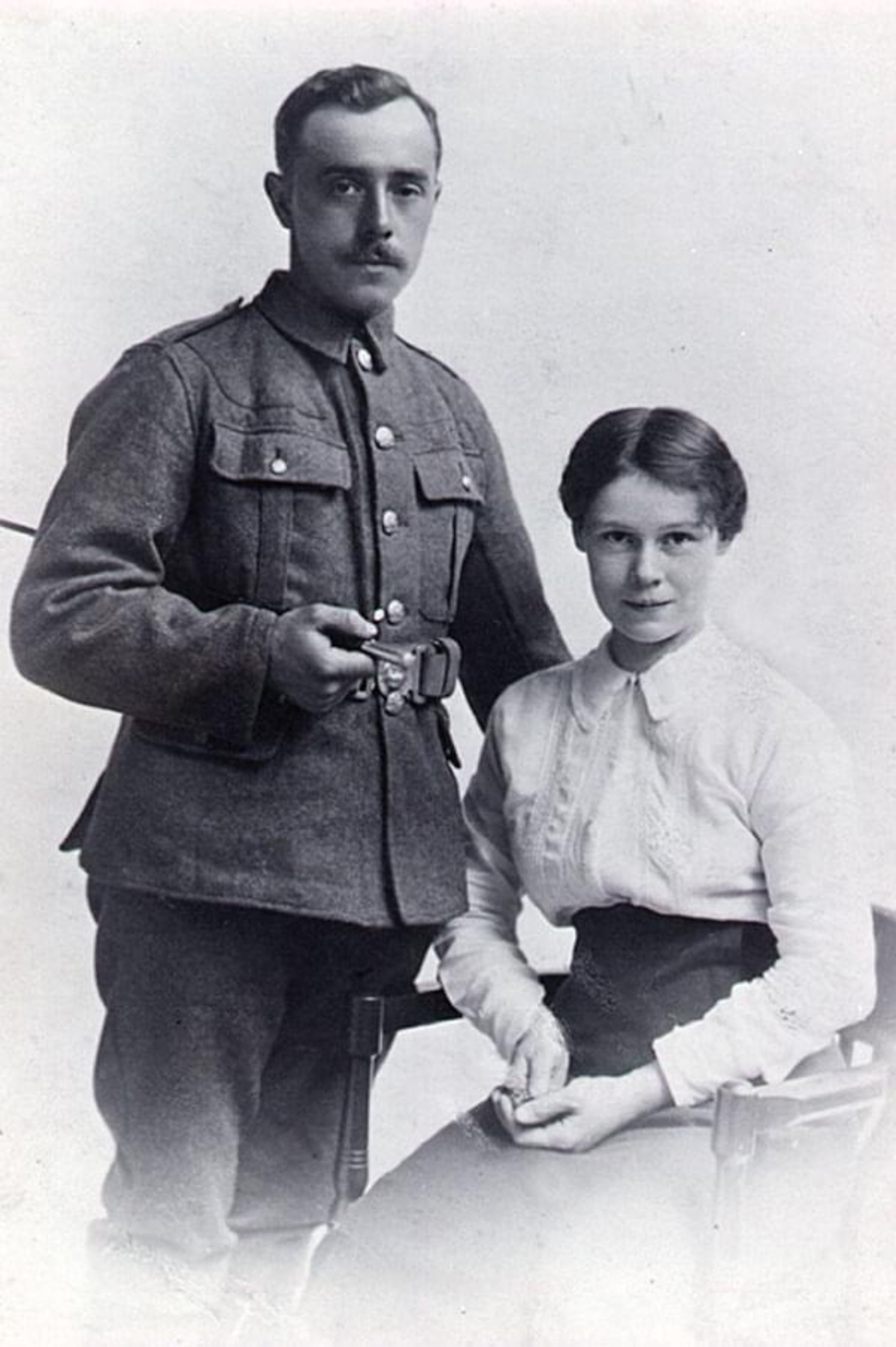 A British Army soldier in uniform and his wife, from the Great War period. He is standing and holding a cane, his wife is seated next to him.