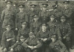 A group photo of fourteen soldiers and one sergeant. The are in three rows in front of a hedge. The front row is seated on the grass.