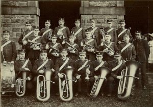 A brass band in uniform, holding their instruments. This is a group of twenty one men arranged in three rows for a group photograph.