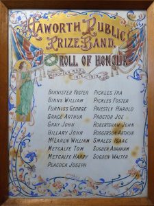 A framed and glazed roll of honour with a thin wooden frame. The colourful decorations at the top have banners, scrolls and flags in full colour and the names are neatly written in two columns. On the left side, a woman in a green dress is holding a laurel wreath over the names.
