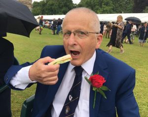 A man in a navy blue suit, shirt and tie about to eat a cucumber sandwich. There is a very long white gazebo in the background.