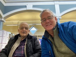 Two older men in a library. They are facing the camera and smiling. In the background the walls are painted blue and cream with white outlines on the arched plaster mouldings and a small diamond shaped window.