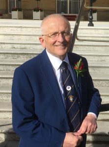 A man wearing a navy blue suit with white shirt and a British Legion tie, standing at the terrace steps of Buckingham Palace. It is a sunny day and he is smiling.