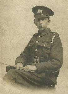 A studio photograph of a soldier in uniform, wearing a service cap with the badge of the West Yorkshire Regiment. He is seated in a chair and on his left shoulder is a light coloured lanyard.