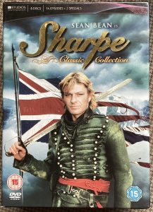 The cover of a DVD boxed set about Richard Sharpe, the hero of the 'Sharpe' series of TV stories starring Sean Bean.It features Sean in uniform dressed as Sharpe, holding a sword with a tattered Union flag flying behind him.
