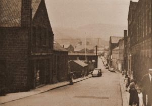 A black and white photograph of Whtiley Street in BIngley, West Yorkshire. There is a road running down the centre of the image, with budilings on either side, many of them are mill buildings of various sizes.