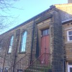 A stone building with arched windows and a brown door with stone steps leading up to it. Clear blue sky above.