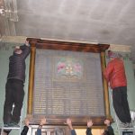 Two men standing on ladders carefully unbolt a war memorial from a wall.