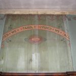 A bare patch behind the memorial shows the Arts and Crafts style decorations previously on the wall. It is a pale green wall with a reddish garland painted on the top just beneath the cornice. There is a curved painted banner with the words 'United We Stand, Divided We Fall.' beneath this is the emblem of the Friendly Society with two hands clasped together in a handshake. At either side can be see a slightly plaer green wall with a different reddish garland painted beneath the cornice.