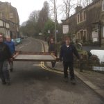 Four men carrying a war memorial down a road. They are passing a sign which says: Belle Isle Road.