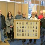 A group of eight people standing around a framed and glazed war memorial. Left to Right. Simon Roarke and Caroline Brown of Keighley Library; Manny and Julie from M & J Framing, Sheila Butler, Tito Arana, Robert Riley and Ian Walkden.