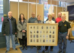 A group of eight people standing around a framed and glazed war memorial. Left to Right: Simon Roarke and Caroline Brown of Keighley Library; Manny and Julie from M & J Framing, Sheila Butler, Tito Arana, Robert Riley and Ian Walkden.