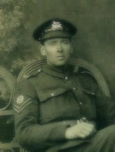 A studio photograph of a soldier in uniform, wearing a service cap with the badge of the West Yorkshire Regiment. He is seated in a chair and on his right upper arm ia a pair of corporal's stripes and above these, a Lewis Gunner proficiency badge showing he was an intructor.