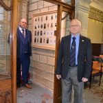 Two men standing either side of a framed and glazed war memorial with photographs of local men.