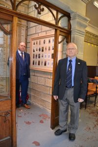 Two men standing either side of a framed and glazed war memorial with photographs of local men.