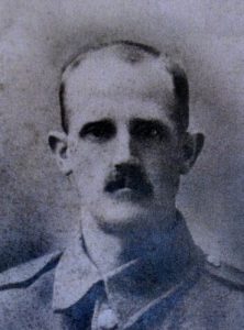 A rather poor quality photo of a man's head and shoulders. He appears to be wearing an army jacker and is facing the camera. He is going thin on top and has a moustache.