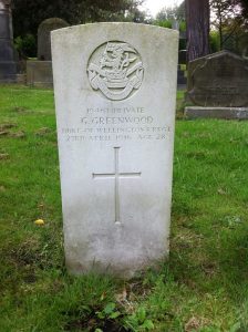 A Commonwealth War Graves Commission headstone. It is rectangular with a curved top and white. Around it are other graves and grass, shrubs and trees.