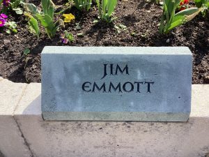 A grey slate stone mounted on top of a low wall surrounding a garden. It bears the name JIM EMMOTT.