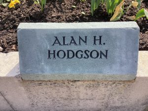 A grey slate stone mounted on top of a low wall surrounding a garden. It bears the name Alan H. Hodgson.