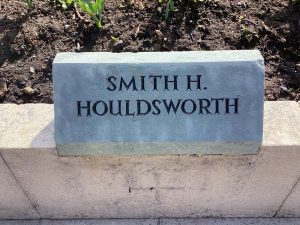 A grey slate stone mounted on top of a low wall surrounding a garden. It bears the name SMITH H. HOULDSWORTH.