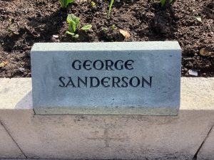 A grey slate stone mounted on top of a low wall surrounding a garden. It bears the name GEORGE SANDERSON.