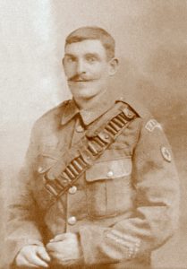 A sepia coloured photograph of a man in British Army uniform. He has a cloth badge on the shoulder with the letters: R.A.M.C. and below this is the Red Cross badge of the Royal Army Medical Corps.
On his lower left sleeve are two chevrons which are good conduct badges. Across his chest is a webbing bandolier.
He has a moustache and is facing the camera.