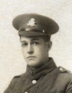A photograph of a West Riding Regiment soldier in a studio. he is wearing a peaked cap with the regimental badge and facing the camera.