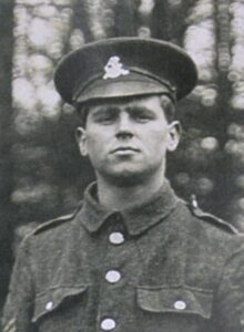 A photograph of a West Riding Regiment soldier in a garden. He is wearing a peaked cap with the regimental badge and facing the camera.