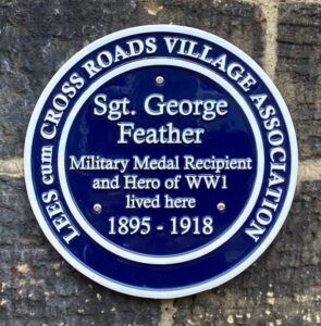 A round blue plaque mounted on a stone wall. The words say: Lees cum Cross Roads Village Association. Sgt George Feather. Military Medal Recipient and Hero of WW1 lived here 1895 - 1918.