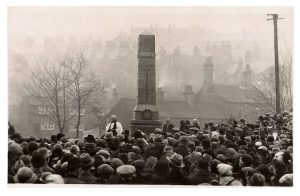 A black and white photograph of a large grey stone obelisk with a stepped base. In the distance is a hillside with houses visible through the mist. Around the memorial is a crowd of people listening to the vicar in the centre.