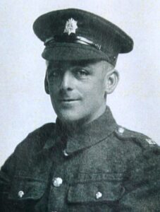 A portrait photo of a British Army soldier in a studio. He is wearing a cap with a regimental badge on it and facing the camera.