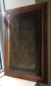 A bronze plaque mounted on a hardwood back mount. It has names in black paint with the initial letters of each word in red paint.
